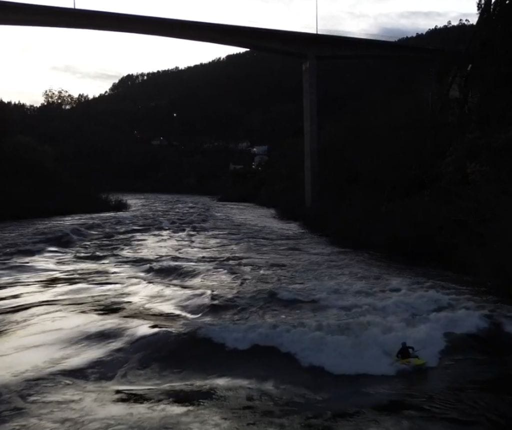 jonathan is seen canoeing in a turbulent river (on the right side) which is surrounded by rocky terrain and a tall and narrow concrete bridge that is spanning the gap between the two shores