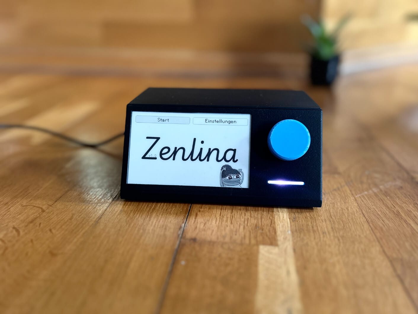 the esp32 e-paper pomodoro timer is pictured here sitting on a wooden desk, a black wire is connected to it, and there is an indoor plant out of focus behind