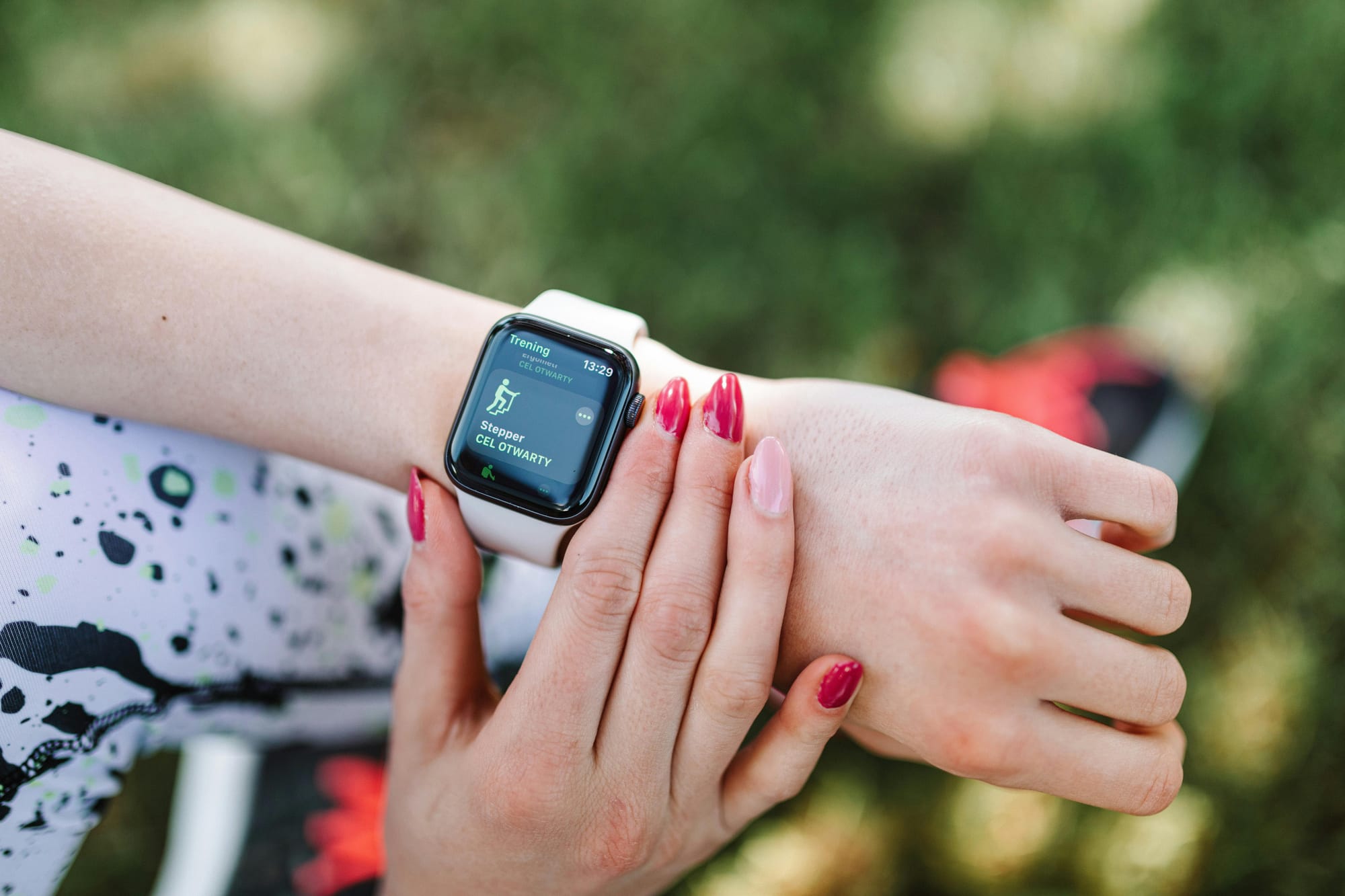 A close up of someone checking their smartwatch with one hand crossed over the other. Green grass in the background, blurred. The visible hand has red nail polish, with the 4th finger having pink nail polish.