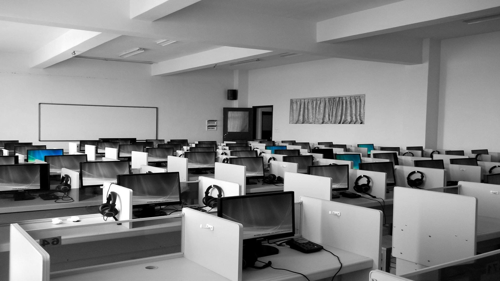 An office setting with white walls and various computer monitors behind different cubicles, alonig with headsets and other peripherals